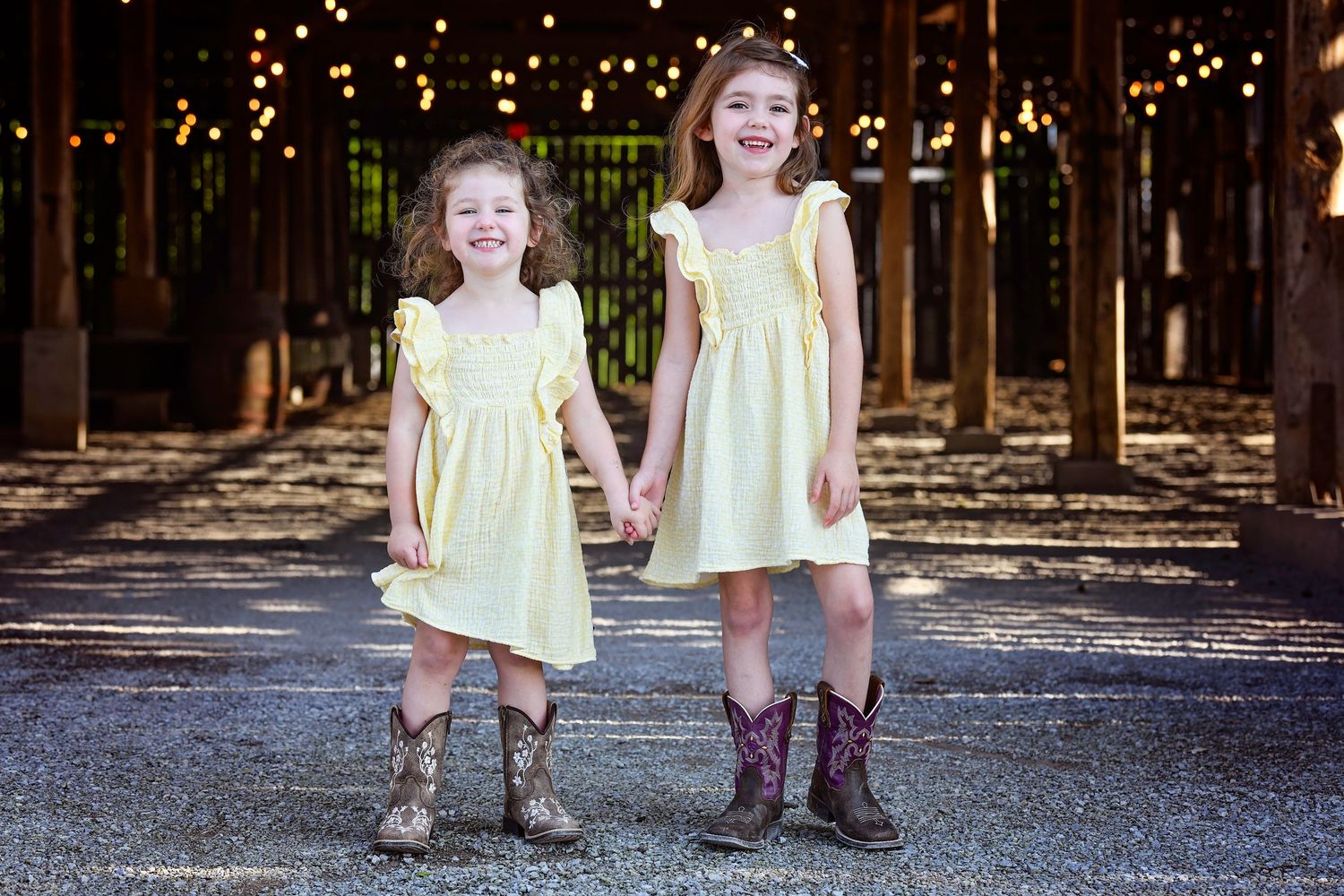 Two children in yellow sundresses and cowboy boots hold hands while standing on a street with string lights above.