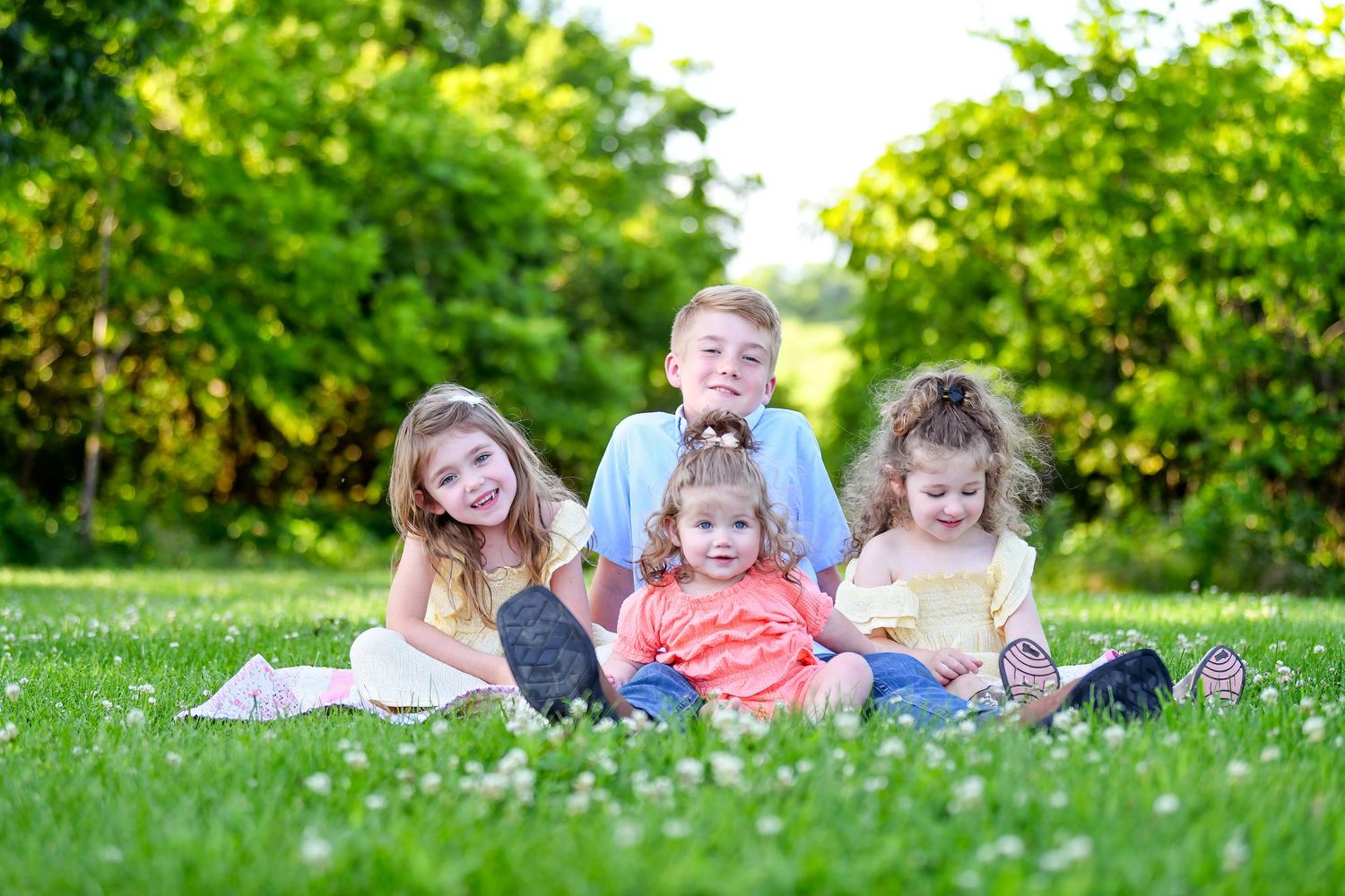 Children sitting together and laughing on green grass in a sunny garden with trees in background.