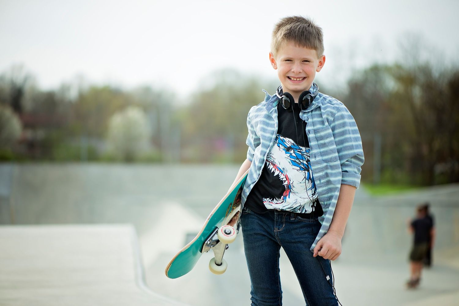 A young skateboarder practices tricks on a paved path while wearing a striped shirt and jeans on an overcast day.