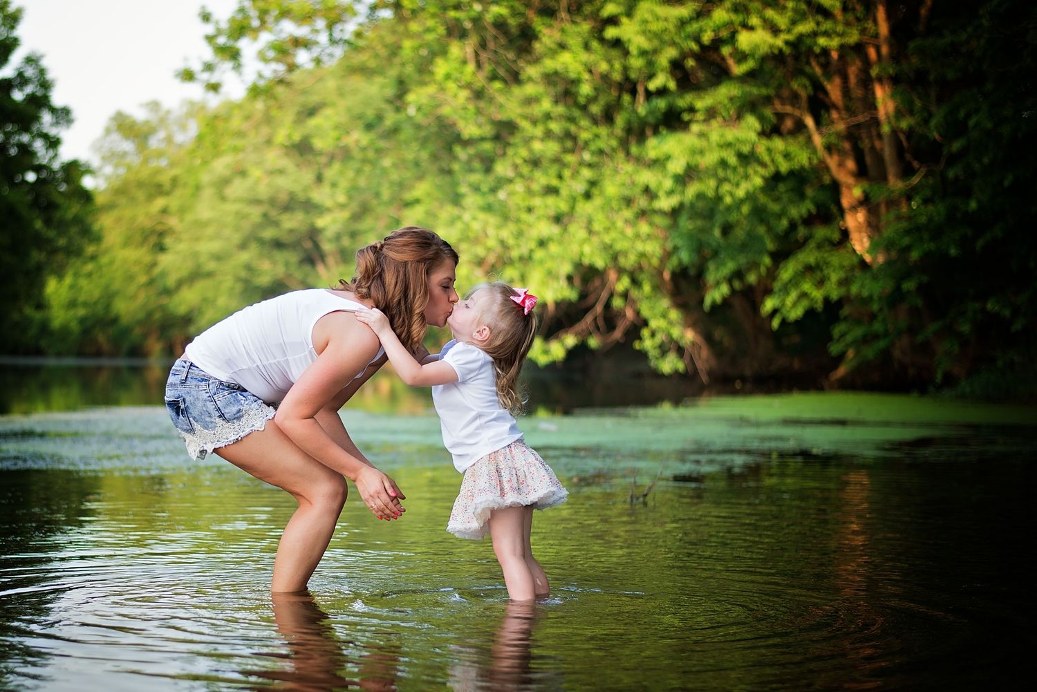 Child splashing in shallow water near lush green trees on a warm summer day.