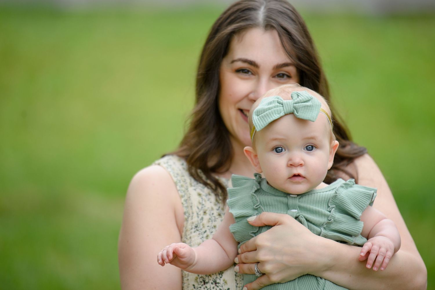 A mother in a floral dress holds her baby girl wearing a sage green outfit and bow headband outdoors.