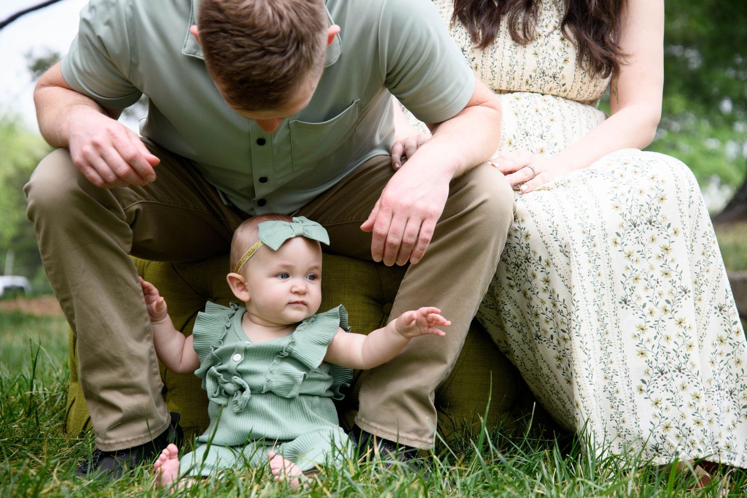 A baby girl in a green outfit sits on grass between her parents during an outdoor family photo session.