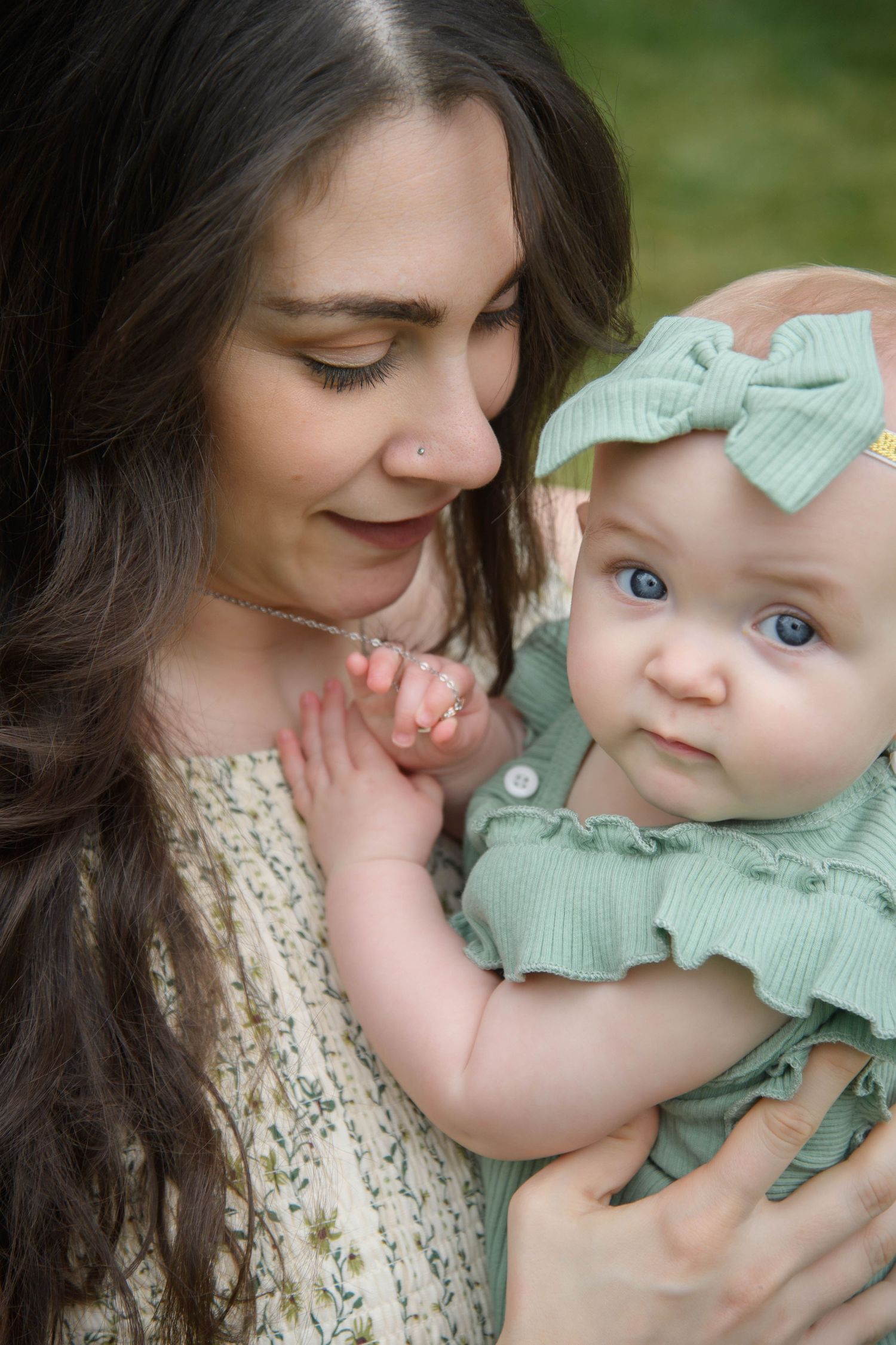 A young mother with dark hair tenderly holds her blue-eyed baby girl dressed in a sage green outfit with matching bow headband.