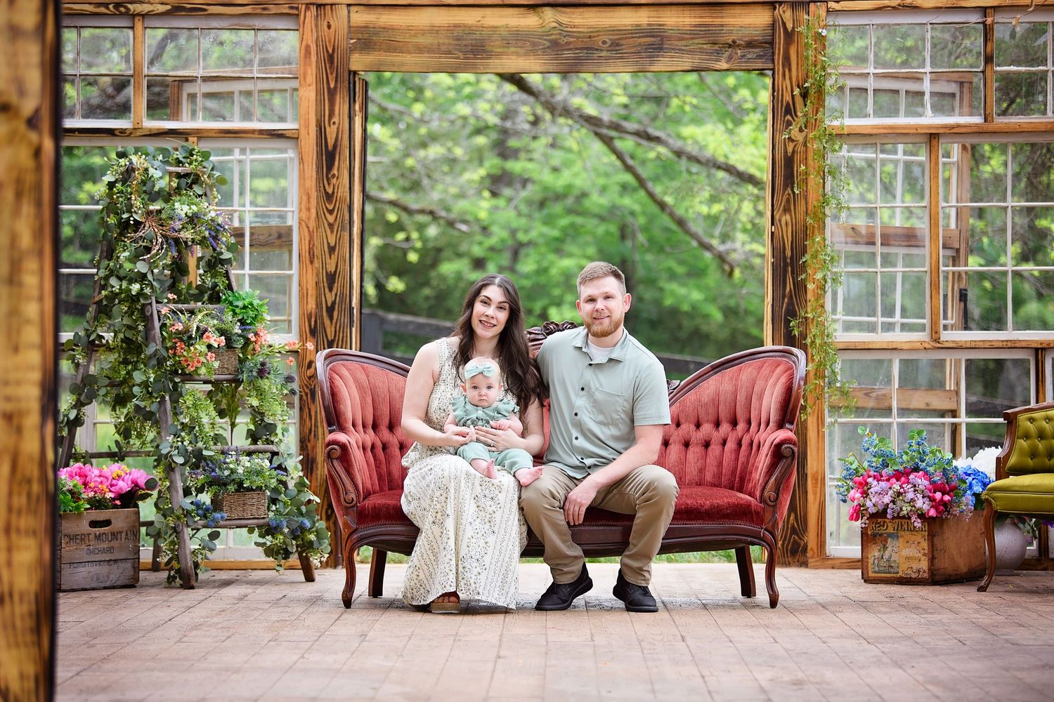 Family portrait on red velvet sofa in rustic greenhouse setting with lush greenery and colorful flowers.