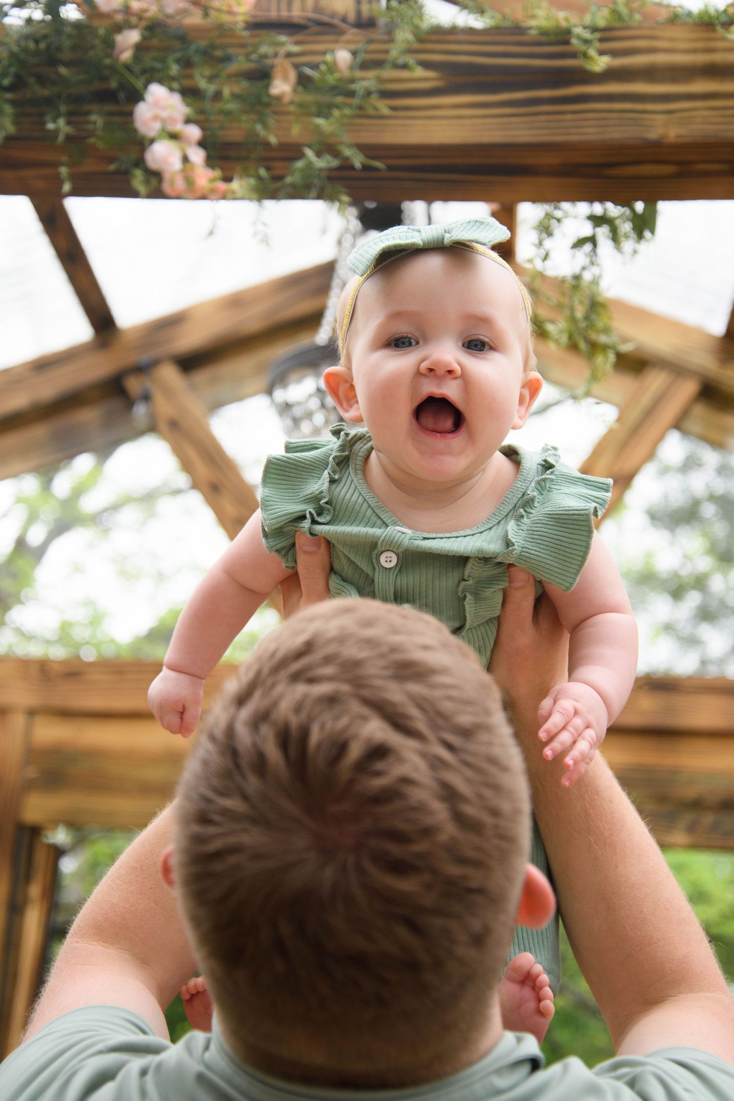 A joyful baby in a green outfit is lifted up by a parent under a wooden pergola decorated with flowers.