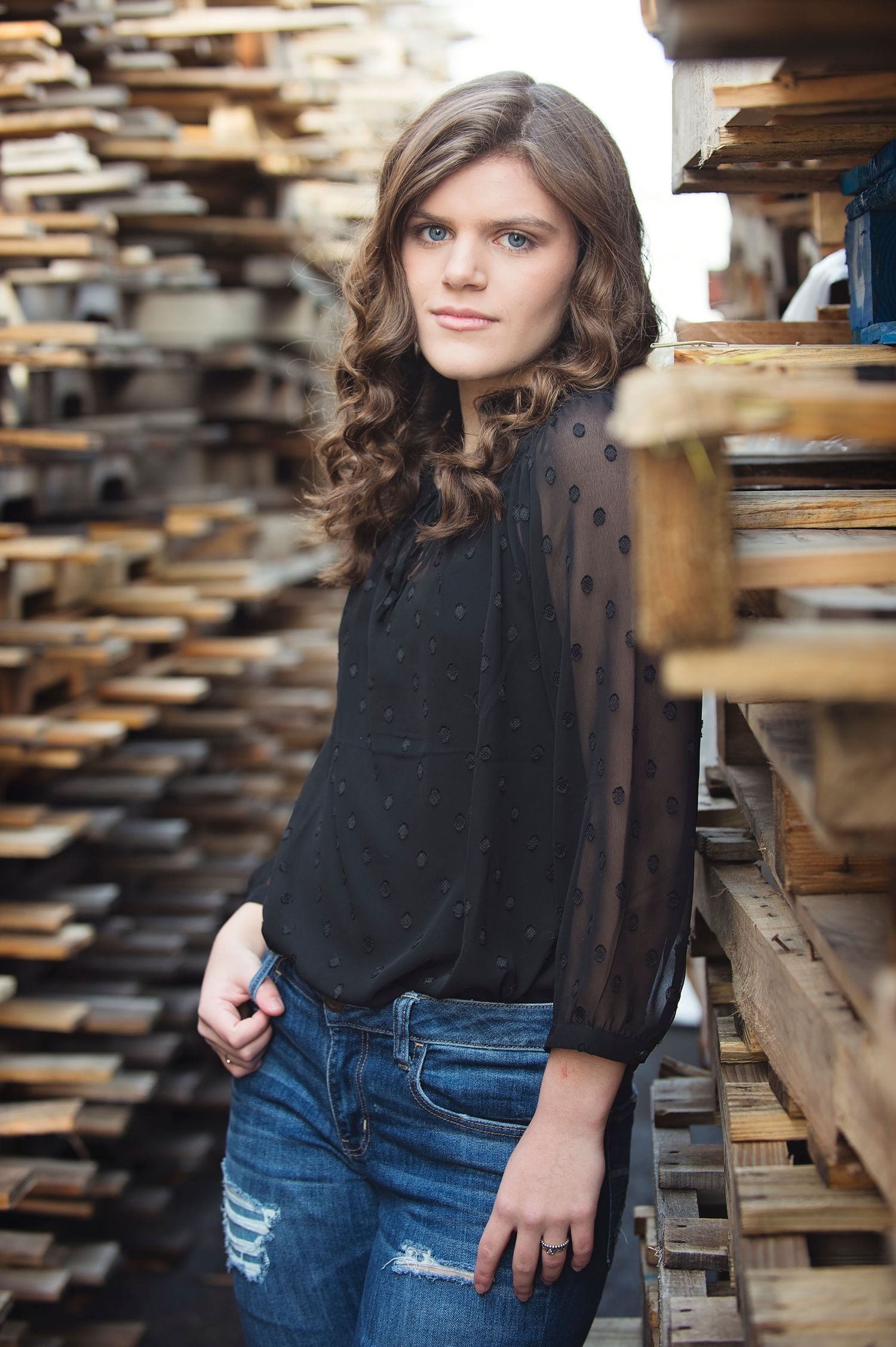 A young person in a black sheer top and distressed jeans poses against wooden pallets in a rustic setting.