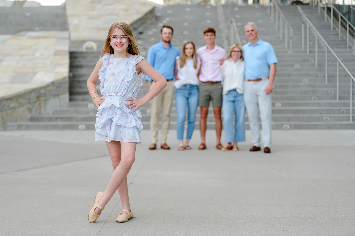 A girl in a blue dress stands in front while a group in casual clothing watches from the steps behind her.