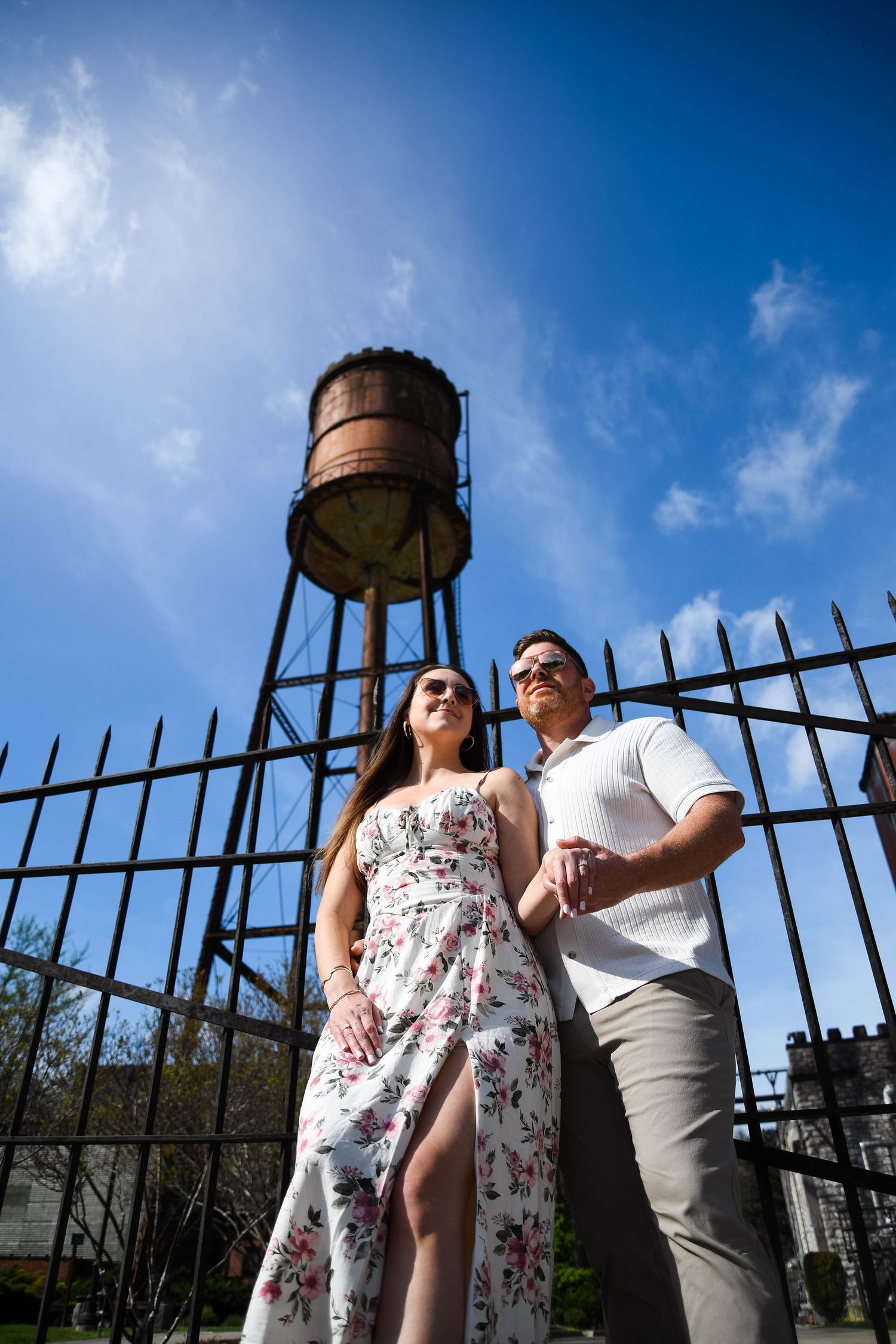 Couple posing in front of rusty water tower and iron fence under bright blue sky, woman in floral dress, man in white shirt.