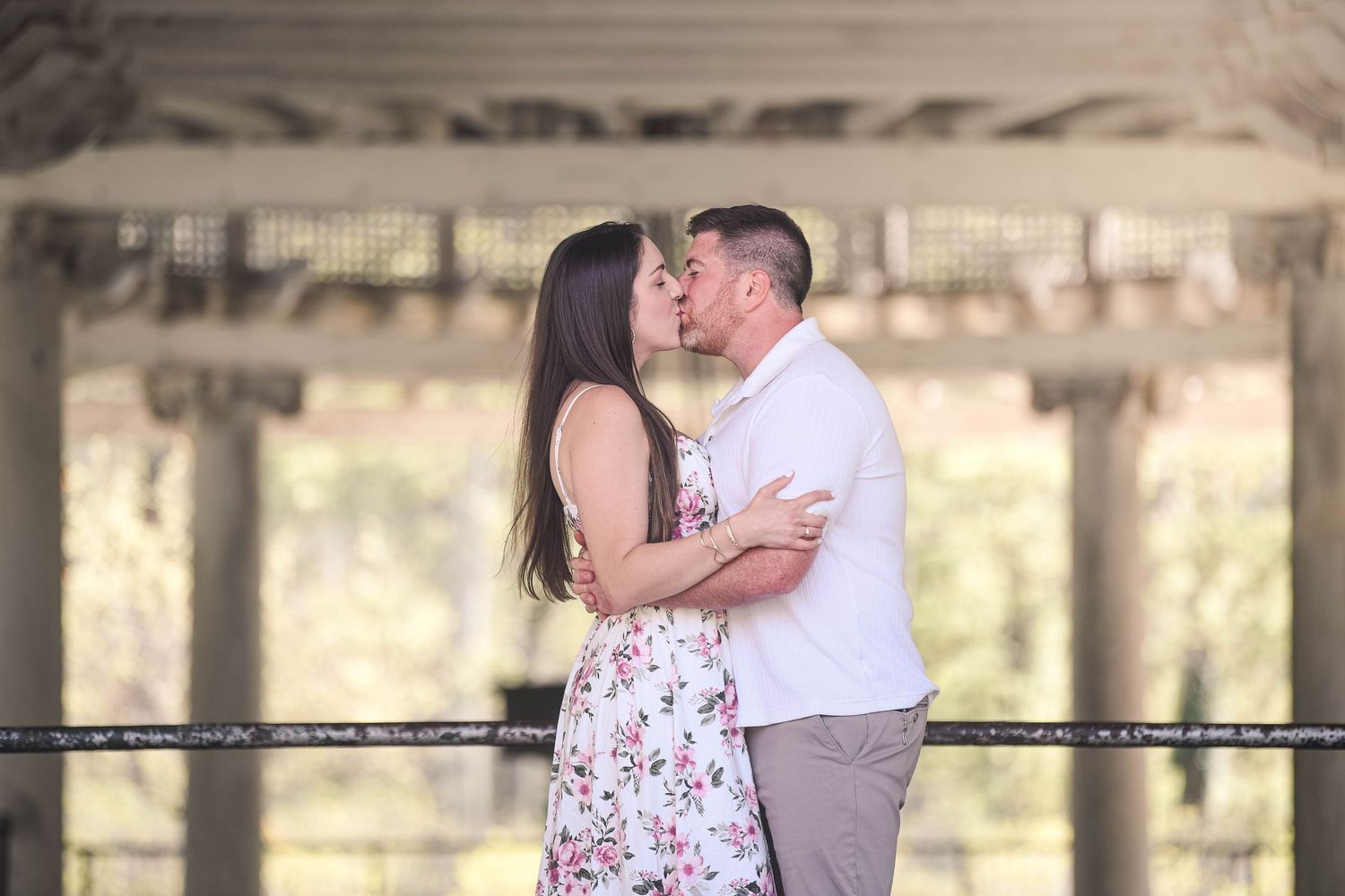 Couple sharing a romantic kiss under a wooden pergola surrounded by lush greenery, woman in floral dress.