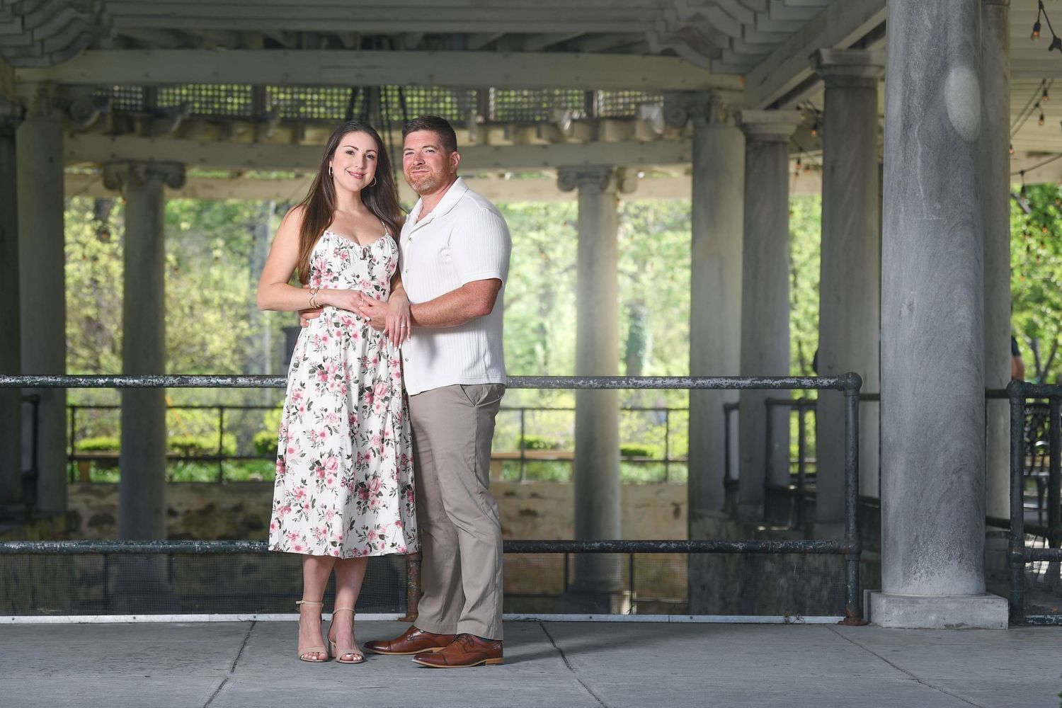 Couple posing together under a pergola in a park, woman in floral dress and man in white shirt, engagement photo.