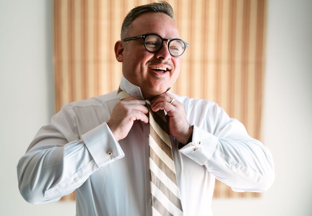 A person in a white dress shirt and striped tie adjusts their bowtie while smiling warmly.