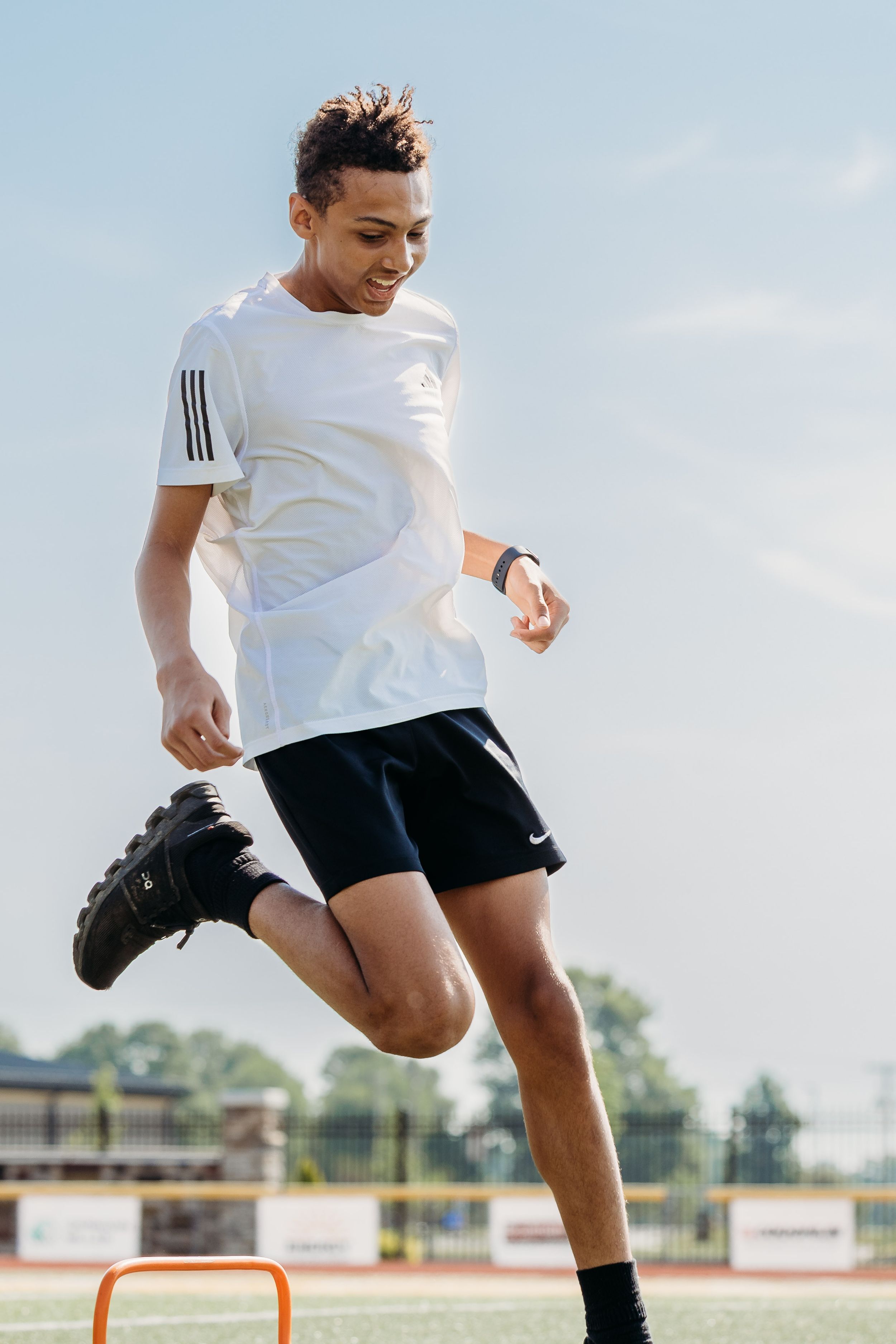 An athlete in white t-shirt and black shorts performs a series of jumping exercises over orange agility hurdles.