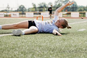 Soccer player lying on artificial turf field after a play with arm raised and goal posts visible in background.