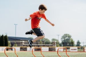 Athlete in red shirt and black shorts jumps over orange hurdles during training session at outdoor sports field.