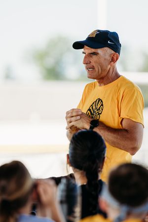 A coach wearing a yellow t-shirt and baseball cap addresses a crowd during an outdoor sporting event.