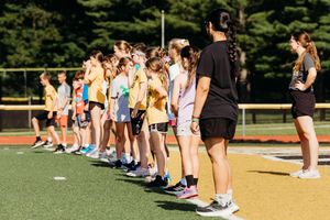 Athletes line up in formation during practice on a sunny outdoor turf field.