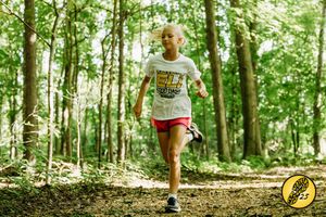 Trail runner wearing red shorts and white tank top jogs through lush summer forest on dirt path.