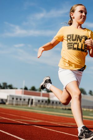 A person wearing an orange t-shirt and white shorts runs on a red track during a sunny day.