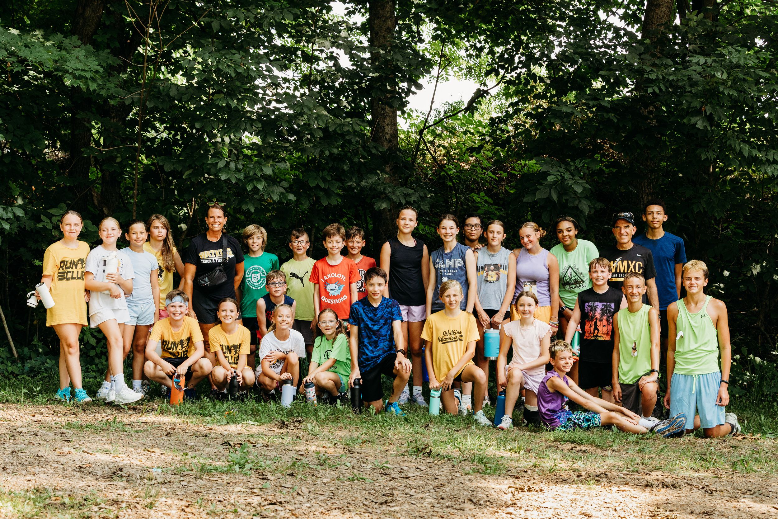 A large group of summer campers and counselors pose together outdoors in casual clothing on a sunny day.