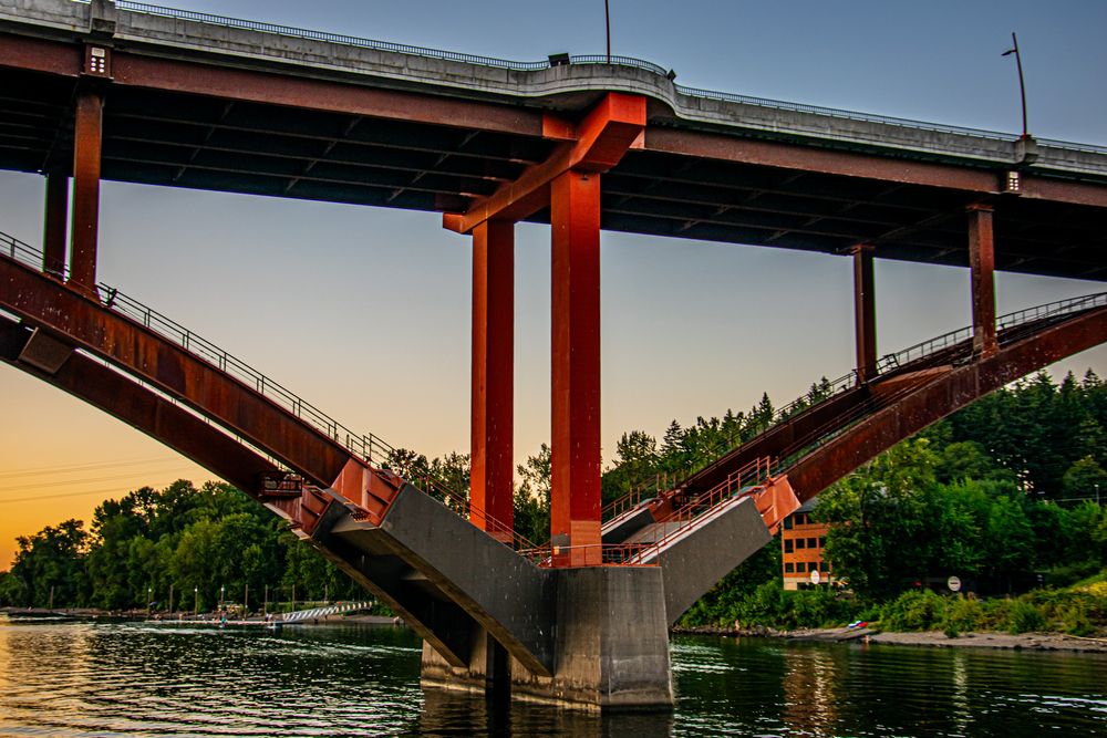 Dramatic red bridge support structure over water at sunset with green trees in background.