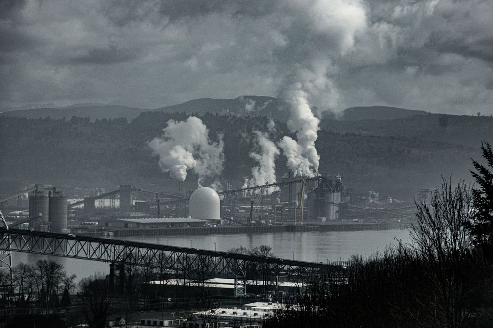 Industrial factory with steam rising against mountain landscape in black and white.