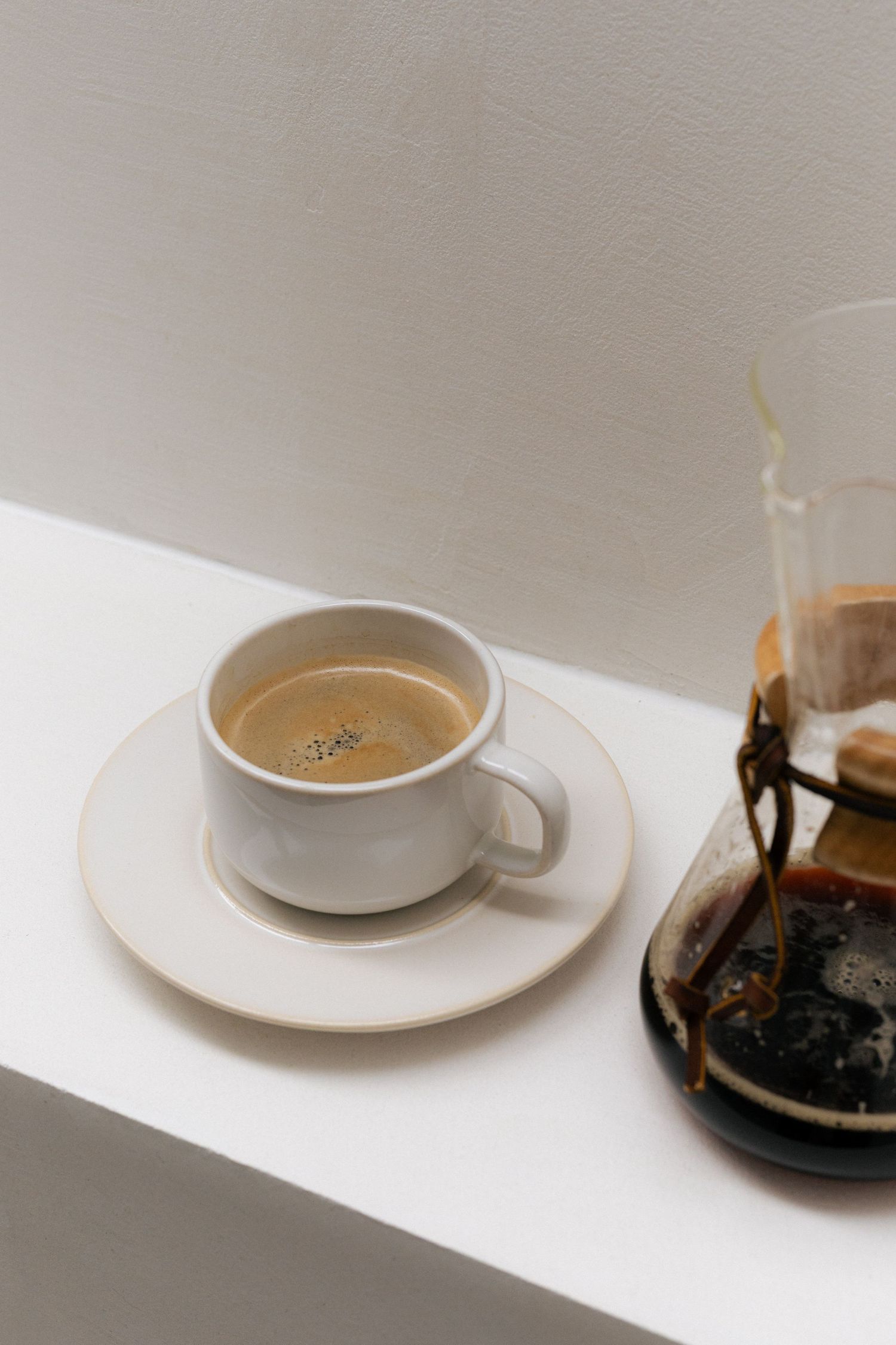 White ceramic coffee cup on saucer beside glass pour-over coffee maker on shelf.