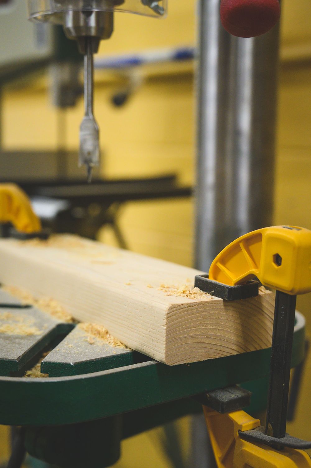 A drill press with cutting bit slowly works through a piece of light-colored wood mounted on a green base.