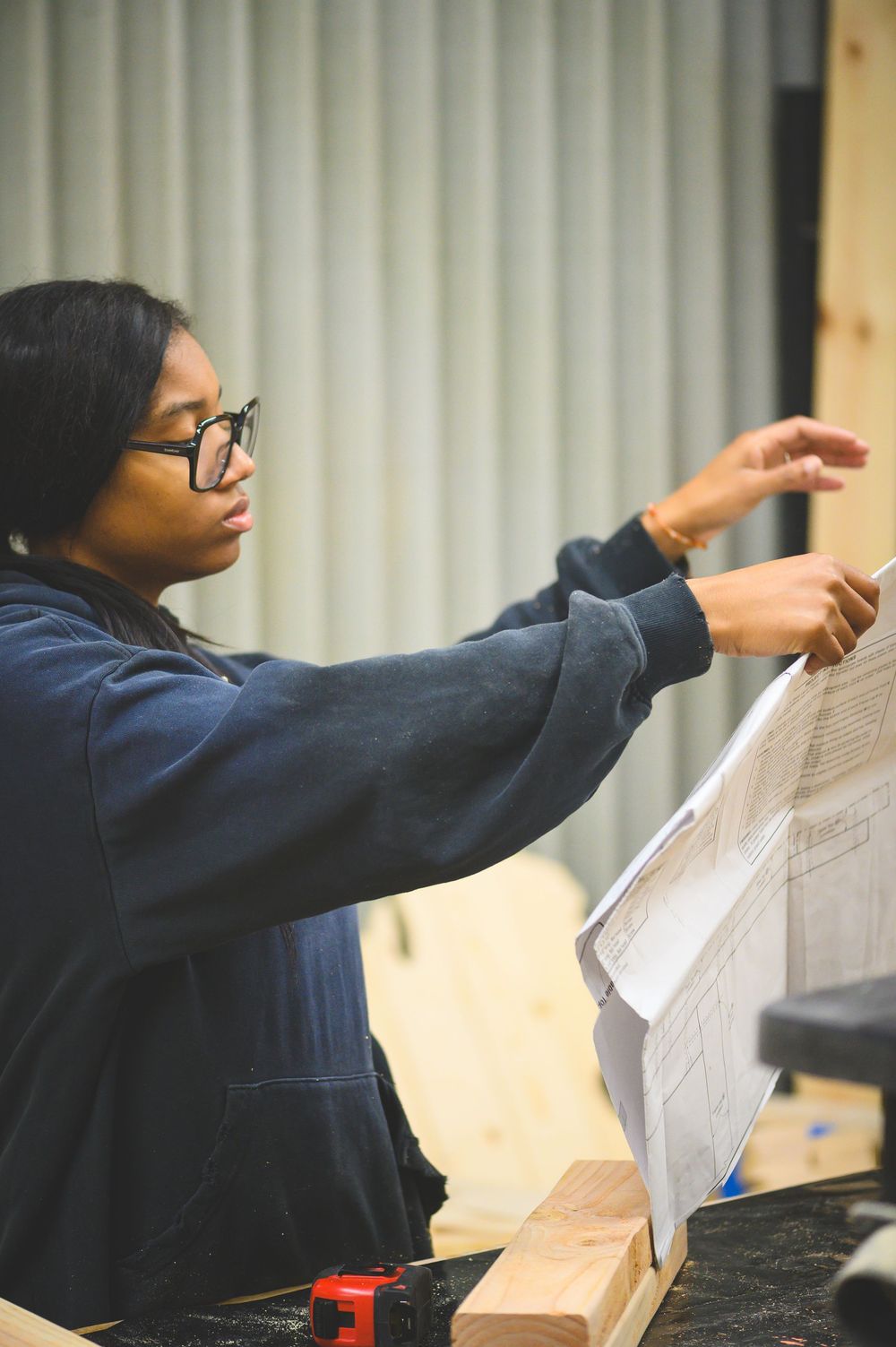 Student in navy hoodie and glasses examines architectural blueprints while working in a workshop setting.