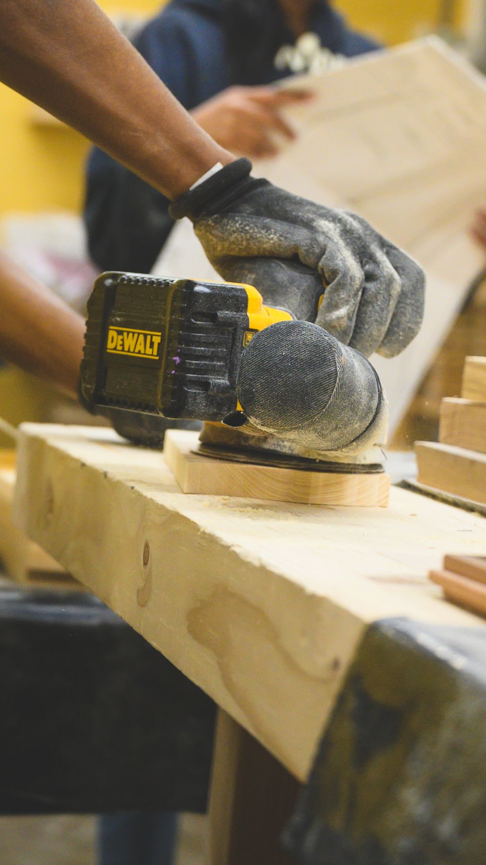 Hands wearing work gloves operate a DeWalt power sander on a wooden surface in a workshop setting.