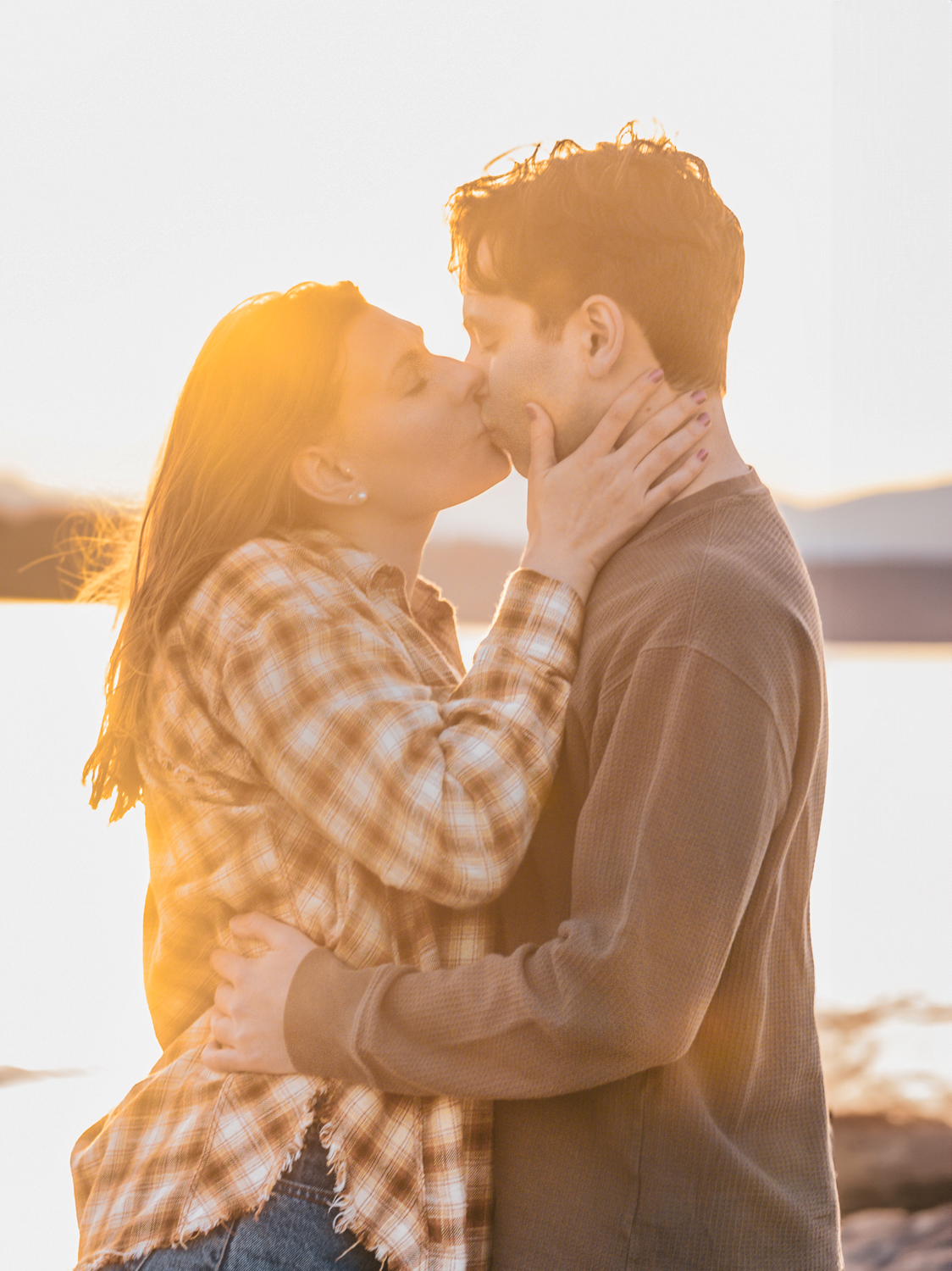 A couple sharing a romantic kiss at sunset by the water while wearing casual autumn attire in golden light.