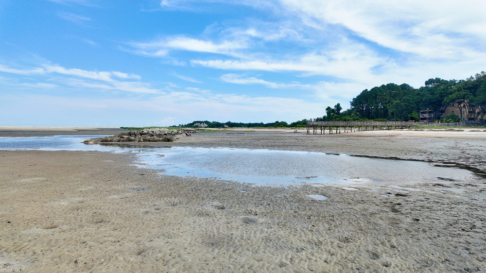 A scenic coastal beach landscape with white sand, shallow waters, and rocky outcroppings under a bright blue sky.