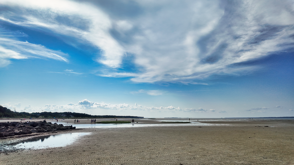 Sweeping sandy beach with dramatic wispy clouds in bright blue sky over calm ocean waters on a clear day.
