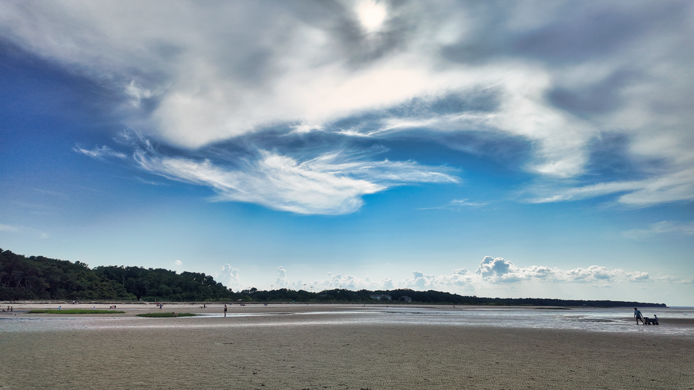 A serene beach landscape with dramatic cloudy skies and distant mountains along a calm shoreline.