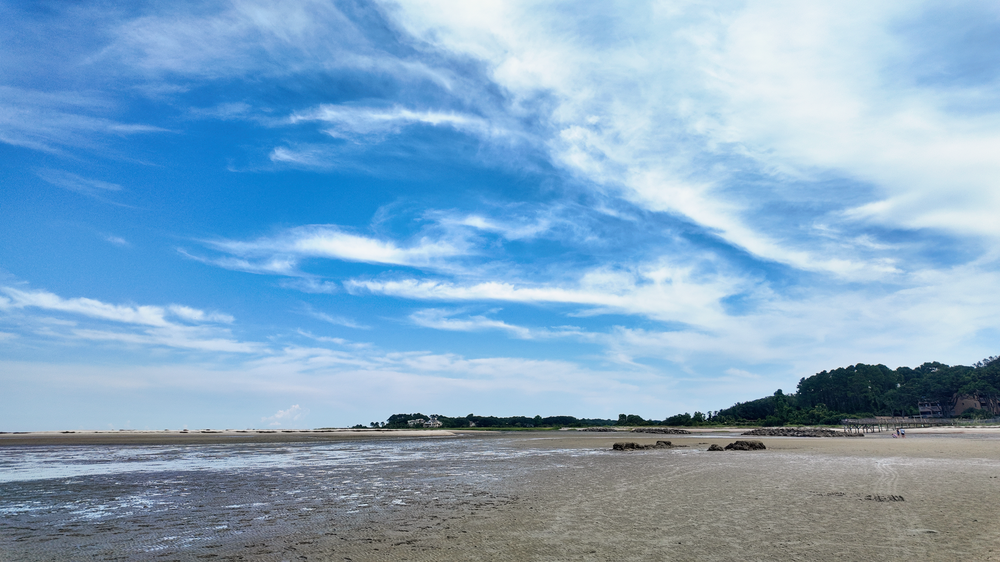 Wide sandy beach stretches along coastline under dramatic wispy clouds and blue sky on a clear day.