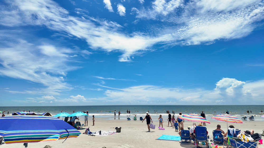 Panoramic view of a sunny beach with colorful umbrellas and beachgoers enjoying the white sand and blue ocean waters.
