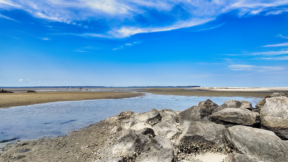 Rocky seawall along a sandy beach with calm waters and bright blue skies with wispy clouds stretching to the horizon.