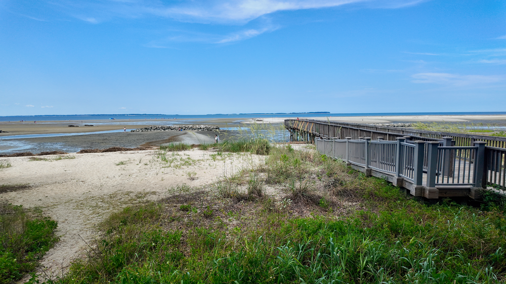 Panoramic vista of coastal beach boardwalk with distant bridge spanning across calm waters and blue skies.