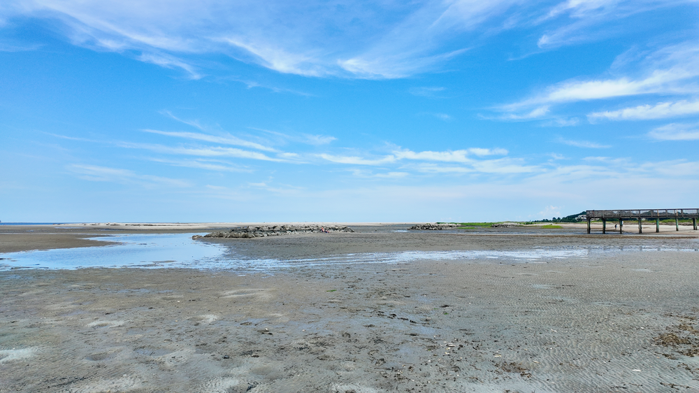 A scenic coastal landscape with mudflats and shallow water under bright blue skies with wispy clouds.