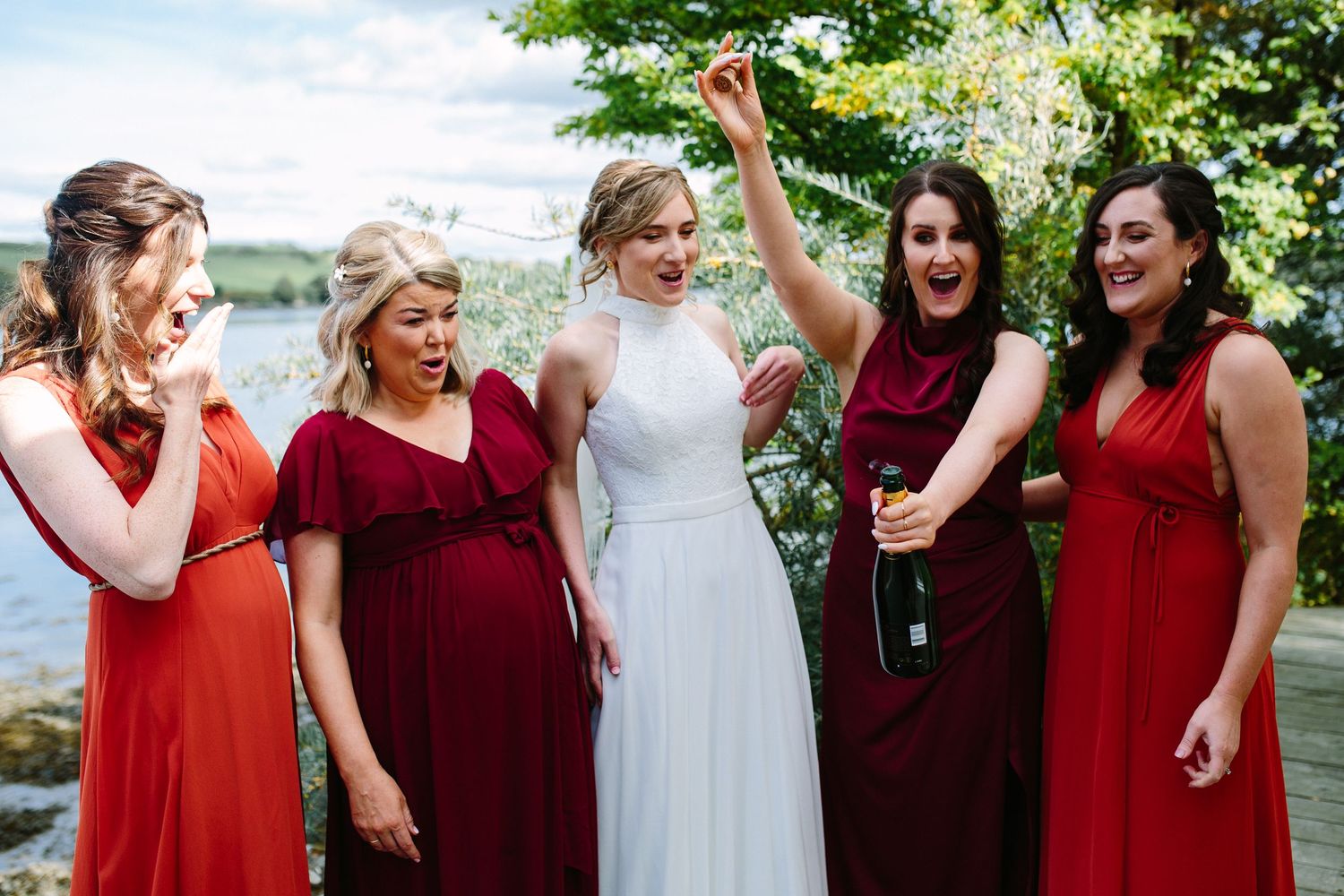 Bridesmaids in burgundy red dresses share joyful candid moments during an outdoor wedding celebration.