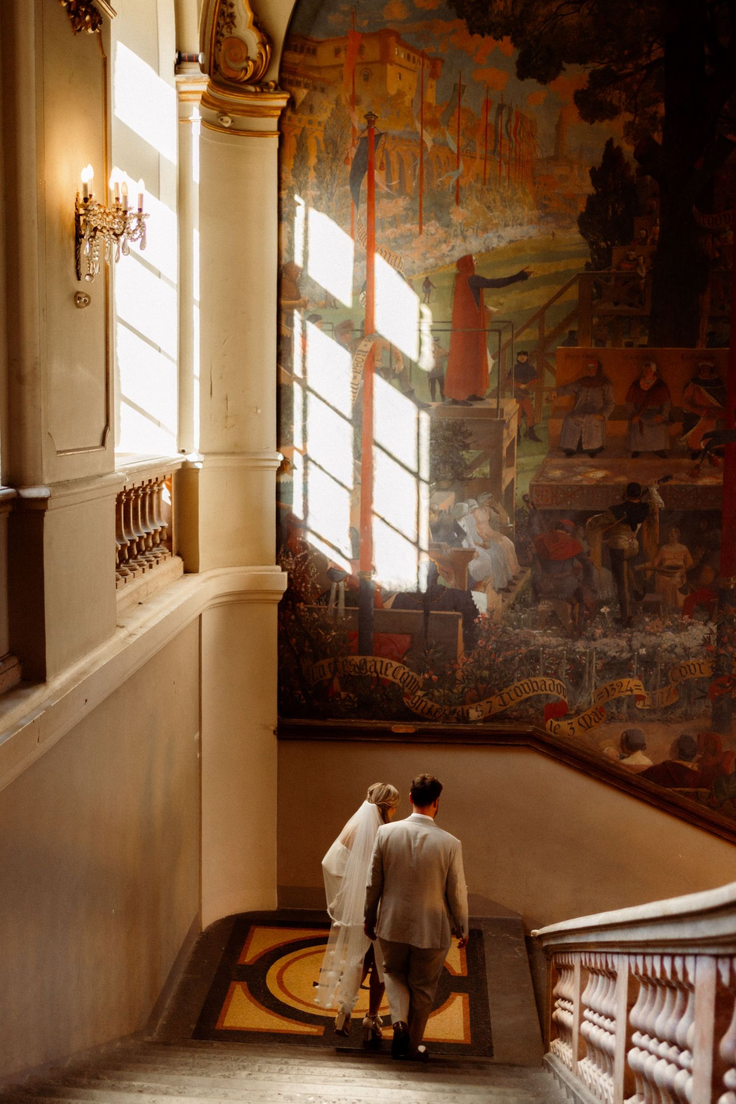 Bride and groom ascending grand staircase beside large mural painting in elegant historic venue.