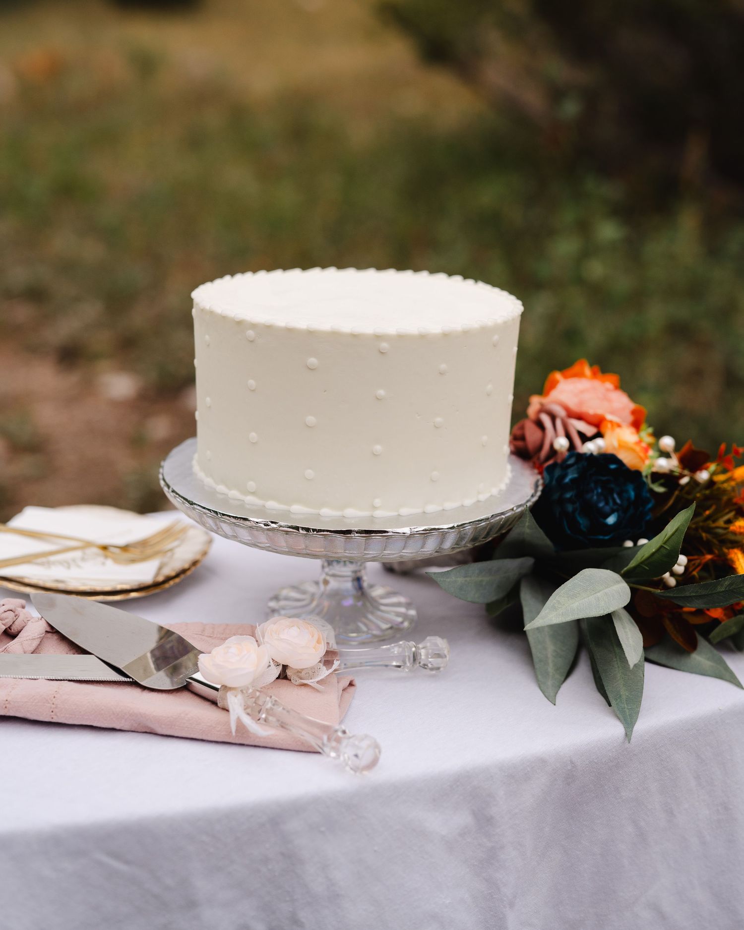 An elegant white wedding cake with swiss dots on a glass pedestal stand, accompanied by floral decor and table settings.