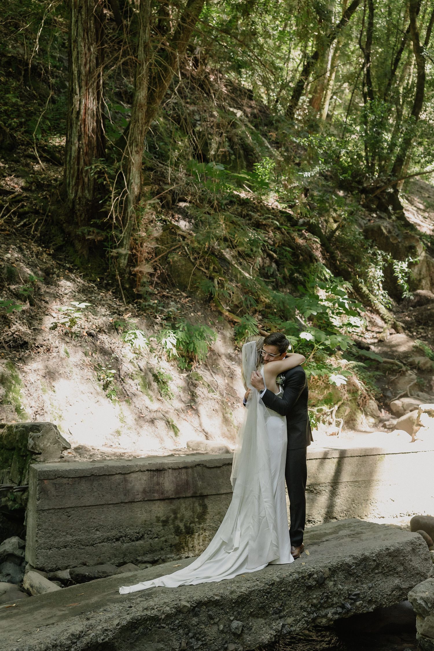 Wedding couple sharing a moment in a lush forest setting with natural stone walls.