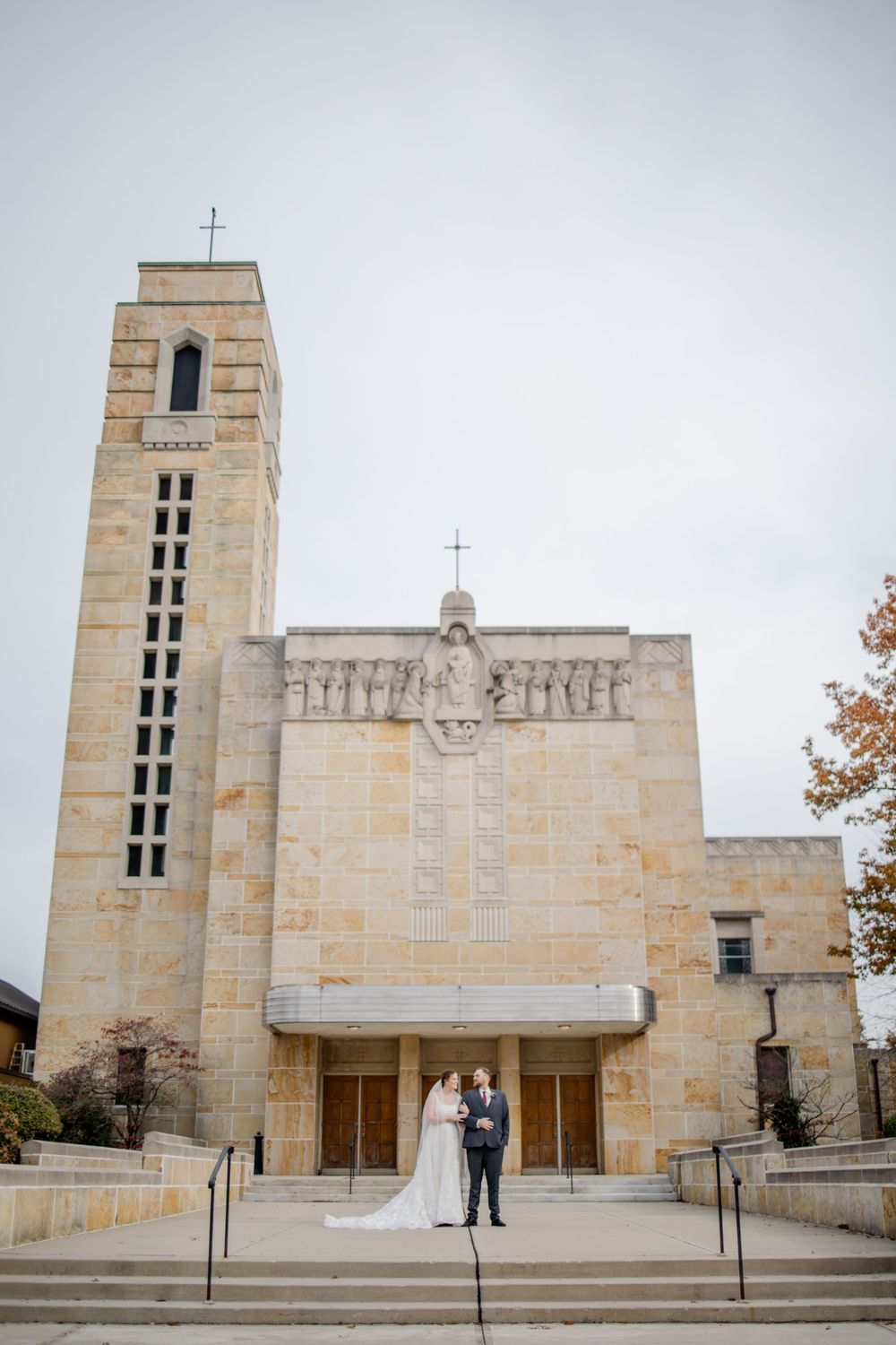A couple stands on the steps of a grand limestone church with a tall bell tower and religious sculptures above the entrance.