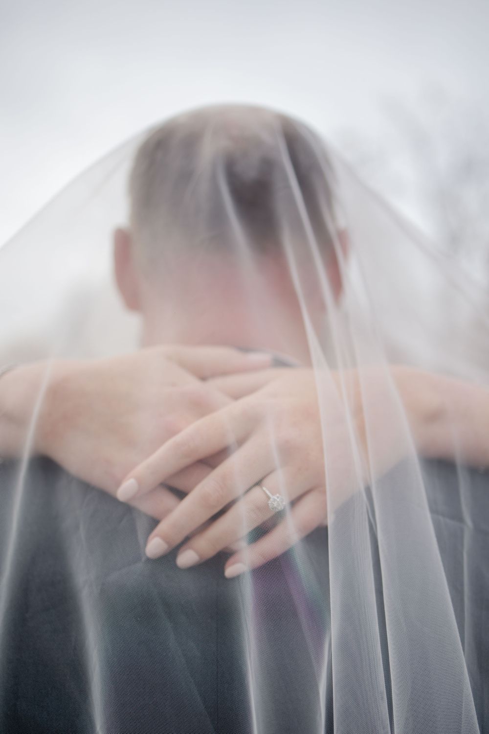 A close-up view of hands under a delicate white wedding veil with soft, ethereal lighting.