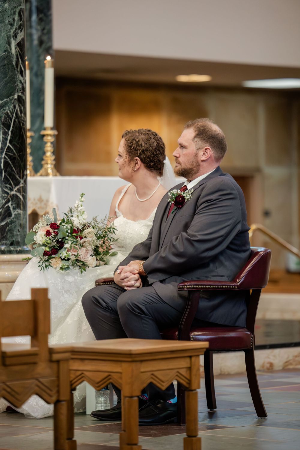 A couple sits together on a bench during a wedding ceremony in a warm indoor setting.