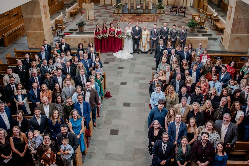 Large group of wedding guests stand together for a panoramic photo in an elegant church interior.