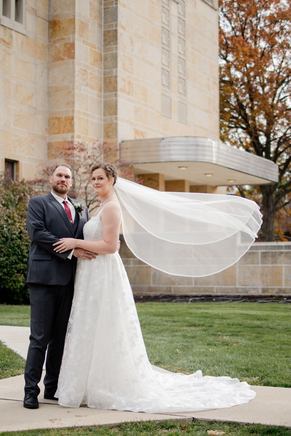A wedding couple poses outside a stone building as the bride's veil flows dramatically in the wind.