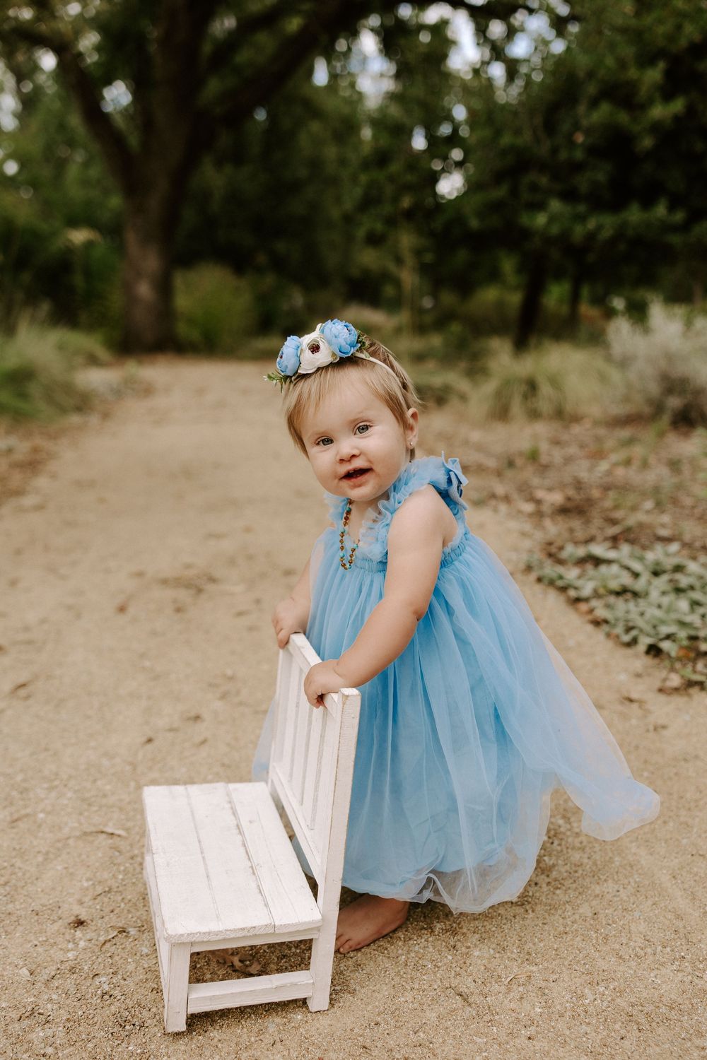 Small figure in a light blue dress and tulle sits on a white wooden bench in an outdoor setting.