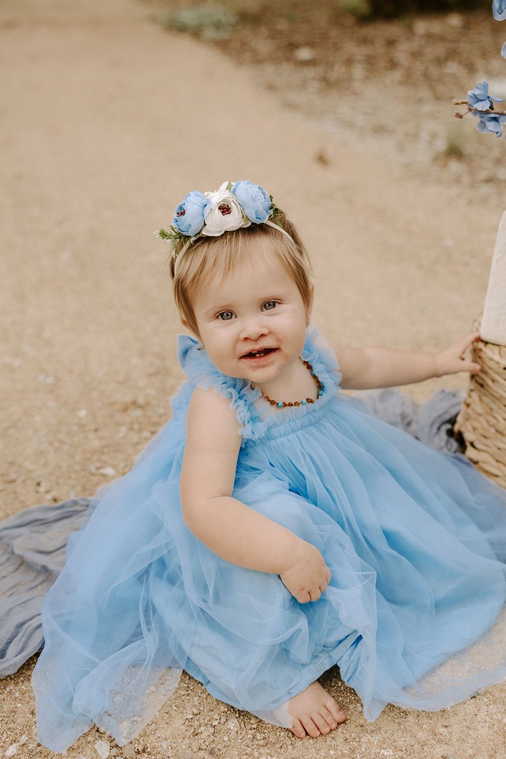 A toddler wearing a light blue tulle dress and floral headband sits on a sandy ground.