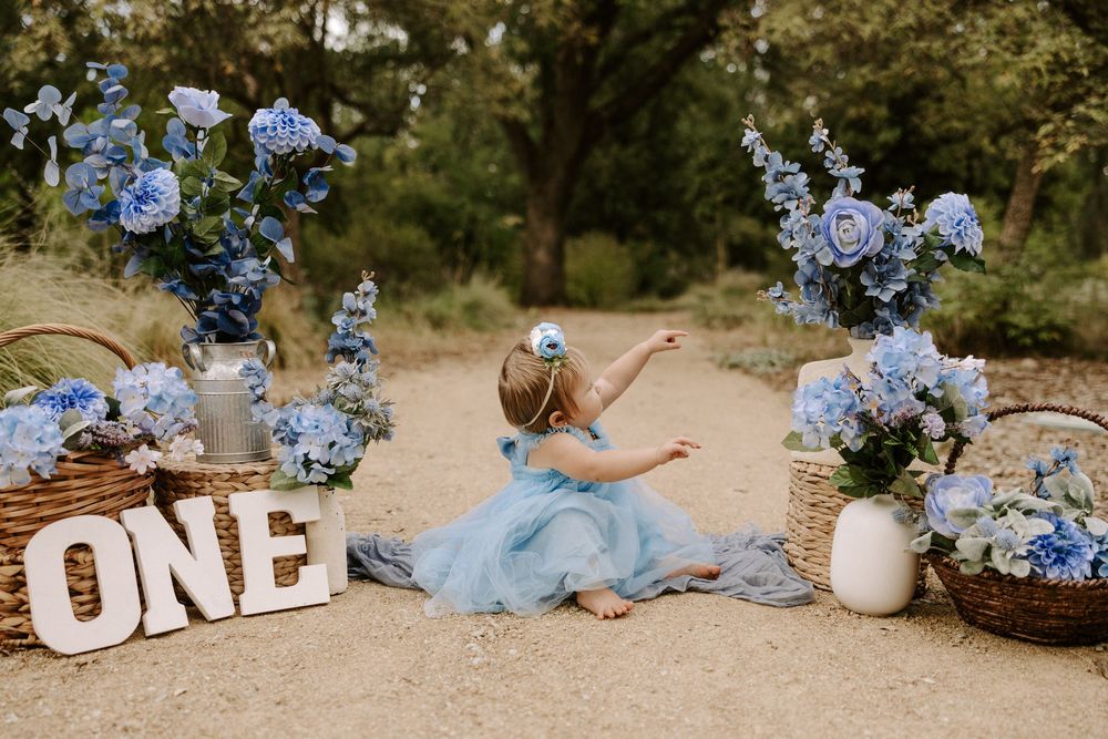 First birthday celebration scene with blue hydrangeas, white 'ONE' letters, and a dreamy outdoor setting.